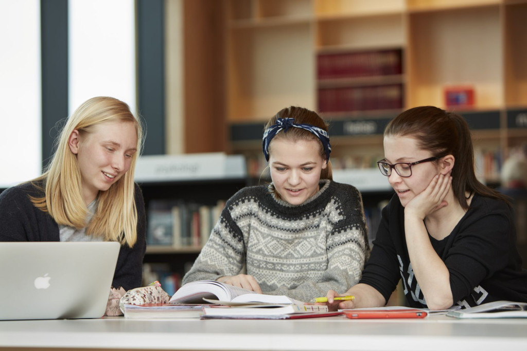 Tre jenter som sitter inne på et bibliotek og jobber med PC og bøker.