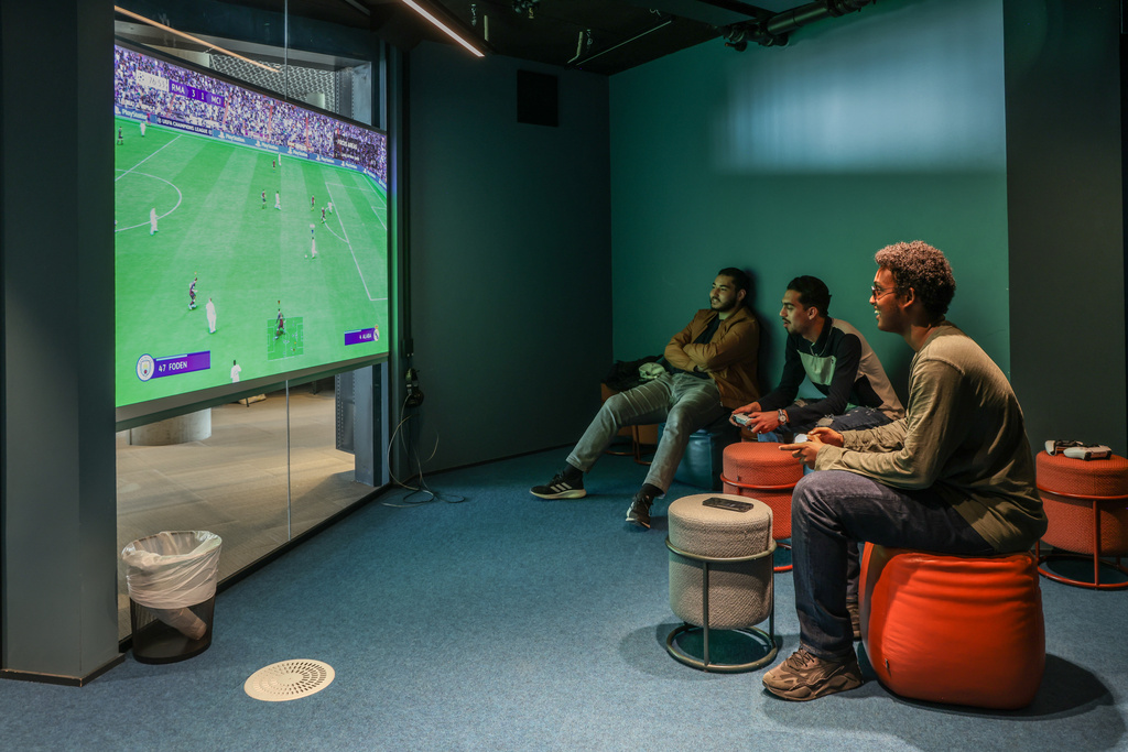 Tre menn sitter i et mørkt rom med stor skjerm og spiller FIFA