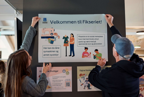 To personer holder opp plakat, venstre kvinne med brunt hår, høyre mann med grått hår og blå caps, på plakaten stård et velkommen til Fikseriet og informasjon om hvordan det fungerer