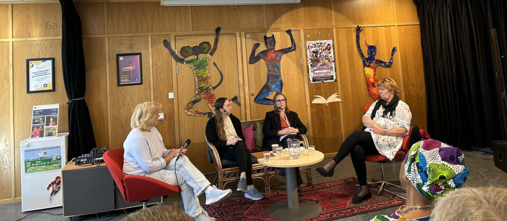 Fire kvinner sitter i sofa og to røde stoler rundt et bord og diskuterer. Fra venstre: kvinne med lyst hår, hvit genser og lys bukse, kvinne med langt brunt hår, lyst topp og svart jakke, mørk bukse, kvinne med brunt hår, briller, burgunder topp og svart jakke, svart bukse, kvinne lengst til høyre, rødt hår, briller på hodet, hvit kjole, svart strømpebukse. Bak innlederne er det ulike figurer på veggen, en som en mus med menneskeklær, en katt i menneskeklær og en okse eller geit med menneskeklær. Plakater henger også på veggen, rødt og svart mønstrete teppe på gulvet.