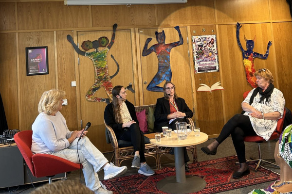 Fire kvinner sitter i sofa og to røde stoler rundt et bord og diskuterer. Fra venstre: kvinne med lyst hår, hvit genser og lys bukse, kvinne med langt brunt hår, lyst topp og svart jakke, mørk bukse, kvinne med brunt hår, briller, burgunder topp og svart jakke, svart bukse, kvinne lengst til høyre, rødt hår, briller på hodet, hvit kjole, svart strømpebukse. Bak innlederne er det ulike figurer på veggen, en som en mus med menneskeklær, en katt i menneskeklær og en okse eller geit med menneskeklær. Plakater henger også på veggen, rødt og svart mønstrete teppe på gulvet.