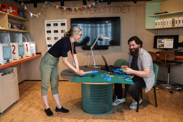 To personer rundt et rundt svart og blågrønt bord. En mann med mørkebrunt hår og skjegg som smiler sitterpå høyre side og ser på et blått og gult verktøy. En smilende litt eldre kvinne med lyst hår i hestehale og briller står til venstre og ser mot mannen. De samarbeider om å bygge noe. PC, lampe og blått byggesett dekker bordet. Bak dem henger en tv, gilander i ulike farger, plakat med instrukser og skilt som sier folkeverksted. Kvinnen har på seg lysegrønn bukse og svart t-skjort og et oransj nøkkelbånd. Svarte joggesko. Mannen har på seg svart bukse og t-skjorte og grå åpen kortskjorte. Hvite joggesko.