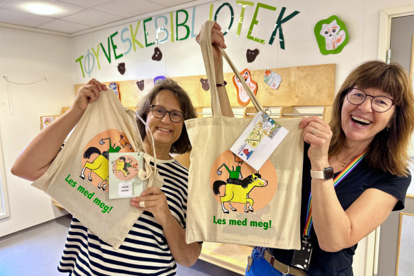 Tøyveskebibliotek. To bibliotekarer holder oppe bærenett med bøker i, på bærenettet står det "Les ,med meg"!