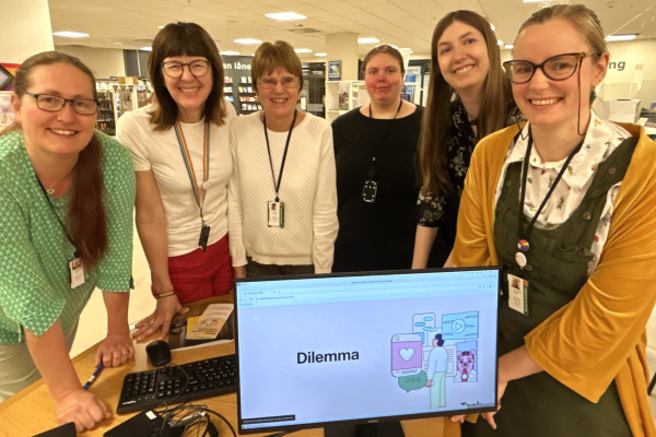 Fem smilende bibliotekansatte står delvis lent foran en skranke i biblioteket. Det står en pc på skranken som viser digital kildebevissthet kurs.