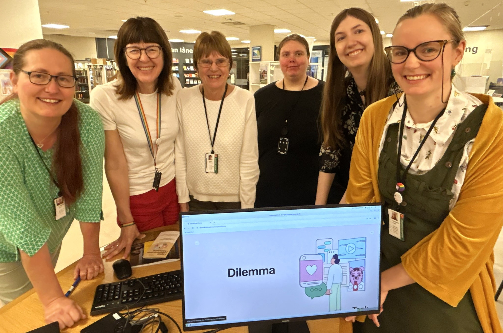 Fem smilende bibliotekansatte står delvis lent foran en skranke i biblioteket. Det står en pc på skranken som viser digital kildebevissthet kurs.