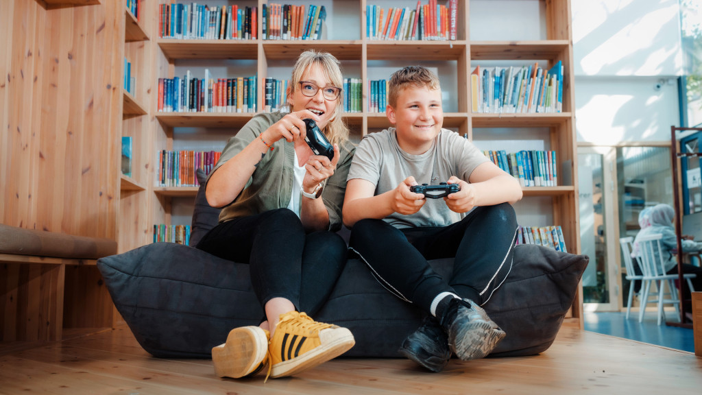 kvinne og gutt sitter på pute på guvlet med spillkonsoller og spiller digitalt spill. Begge smiler.
