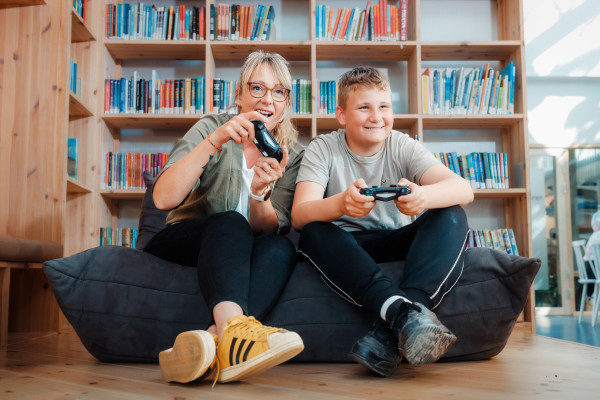 kvinne og gutt sitter på pute på guvlet med spillkonsoller og spiller digitalt spill. Begge smiler.
