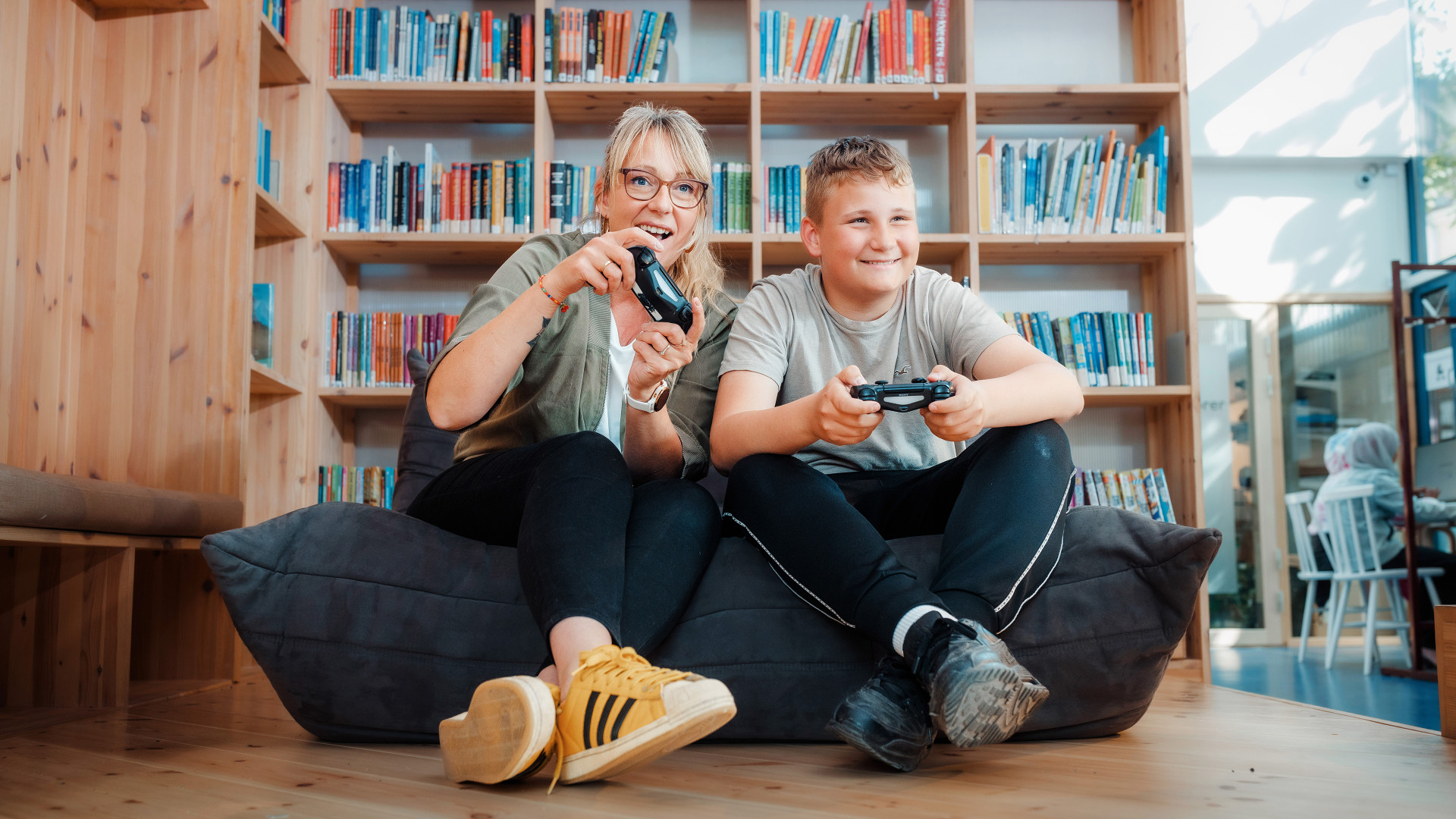 kvinne og gutt sitter på pute på guvlet med spillkonsoller og spiller digitalt spill. Begge smiler.