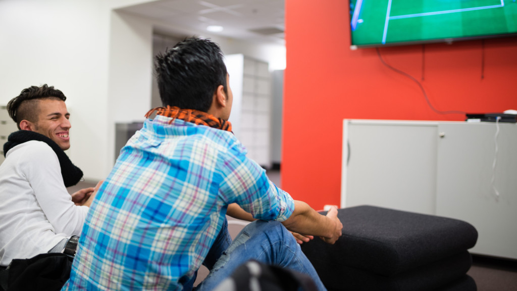 To ungdomsgutter ser mot TV-skjerm med spillkonsoll i hendene og spiller digitalt spill. Den ene smiler mens den andre ser man bakhodet til