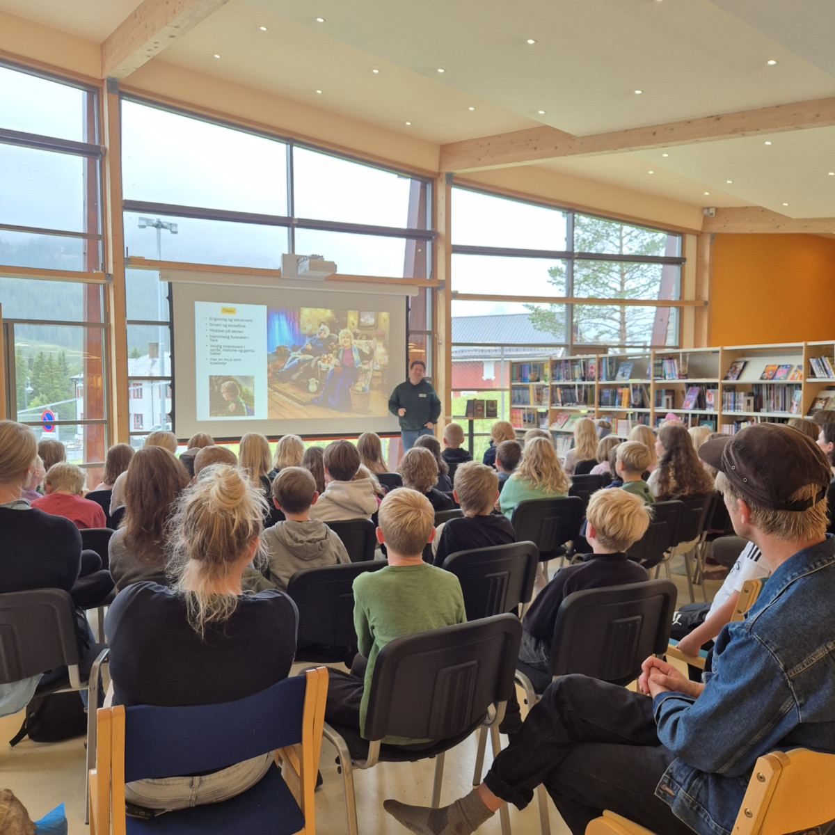 forsamling med barn i bibliotek som ser mot bibliotekansatt som formidler foran lerret