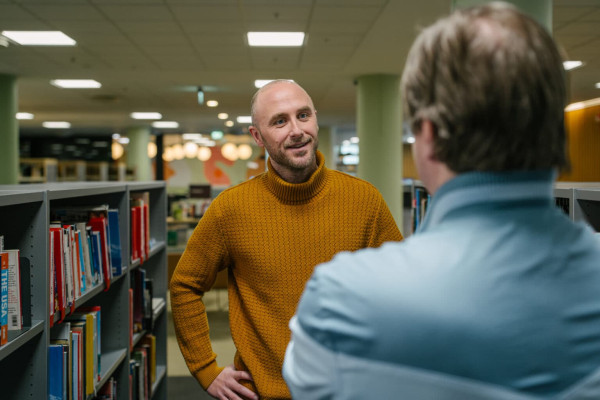 to menn står blant bokhyllene i et bibliotek. Den ene i blå skjorte med ryggen til kamera den andre i burgunder-brun genser ser mot den andre som smiler litt, fjeset synlig i kameravinkelen