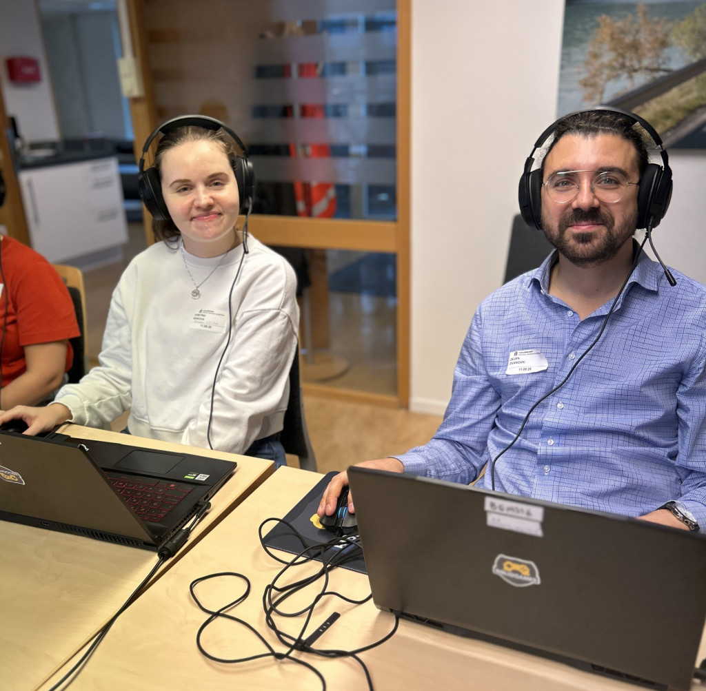 ung voksen kvinne i grå genser og mann i blå skjorte og briller sitter ved siden av hverandre med headset og datamaskiner og smiler mot kamera