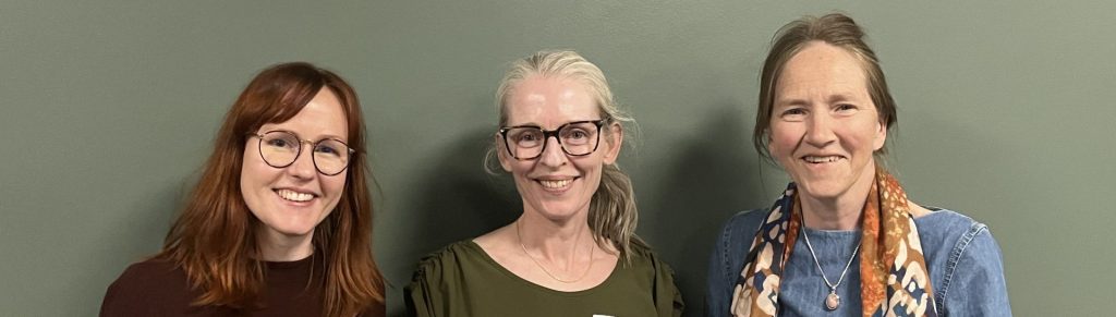 Tre kvinner står ved siden av hverandre, ser mot kamera og smiler. De to første har briller.